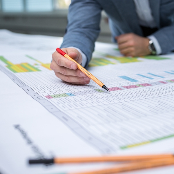 Eine Person in einem Anzug schreibt mit einem orangefarbenen Stift auf einem großen, farbig markierten Tabellenblatt.