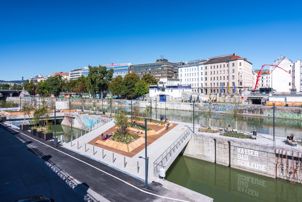 Städtische Uferpromenade mit Sitzbereichen und Bäumen am Fluss, von Gebäuden und Graffiti-Wänden umgeben, bei klarem Himmel.