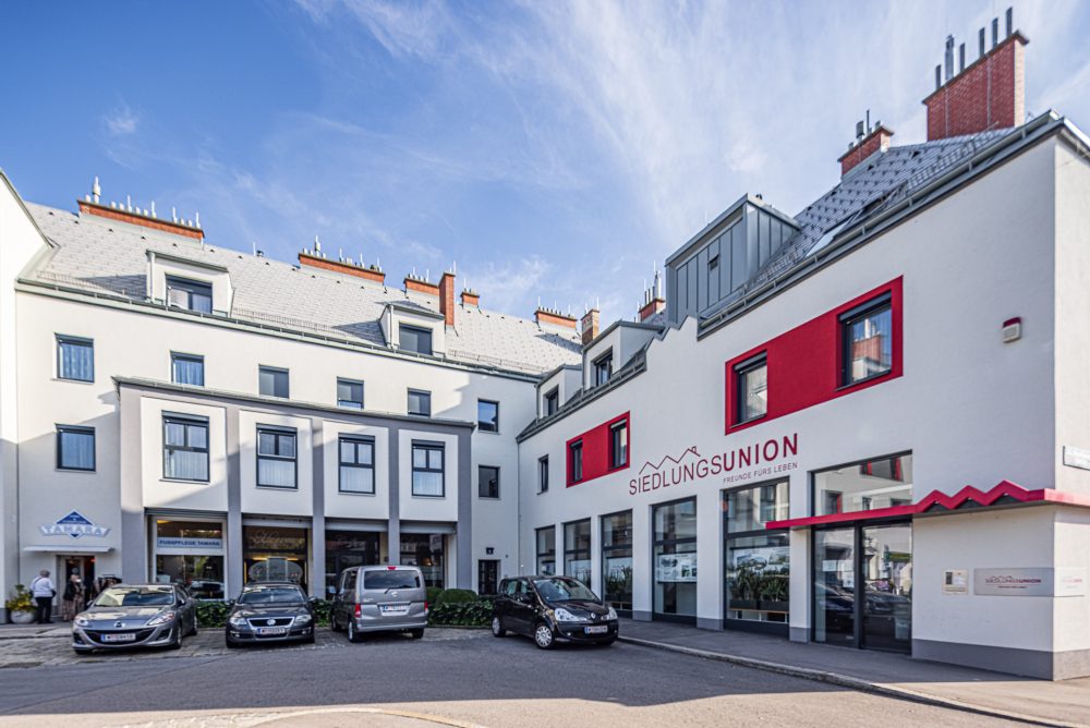 Das Bild zeigt ein modernes Bürogebäude mit der Aufschrift "Siedlungsunion" und mehreren geparkten Autos davor, bei klarem Himmel.