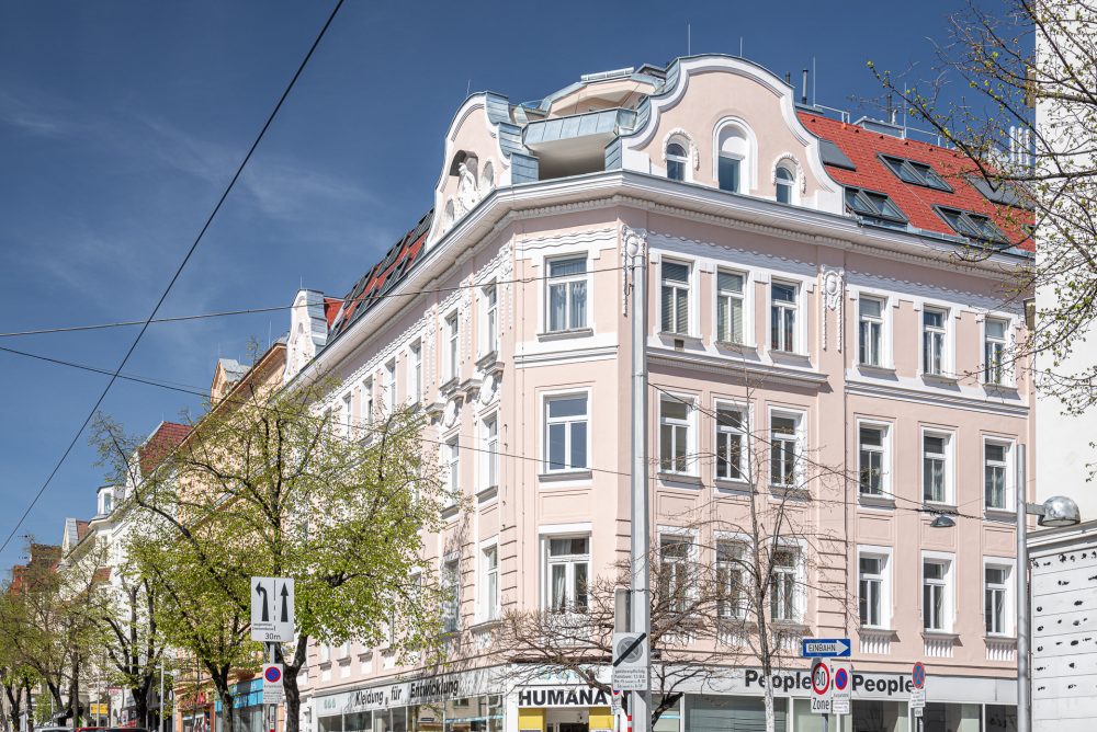 Ein rosa-weißes historisches Gebäude an einer Straßenecke mit mehreren Etagen und Geschäften im Erdgeschoss unter klarem, blauem Himmel.