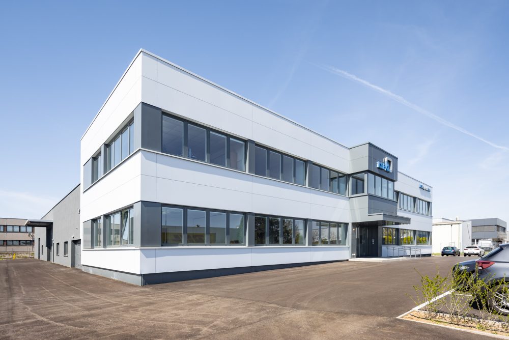 Modernes, zweistöckiges Bürogebäude mit weißen Paneelen, großen Glasfenstern und einem Parkplatz im Vordergrund an einem klaren Tag.