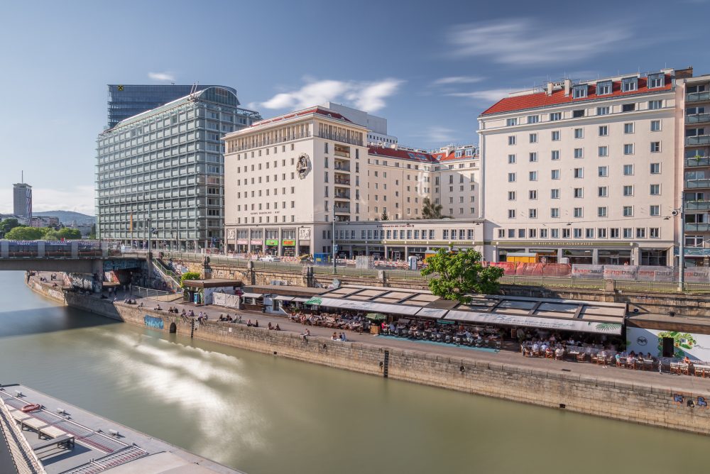 Stadtansicht mit modernen und traditionellen Gebäuden entlang eines Flusses, Menschen sitzen am Ufer unter einer überdachten Terrasse.