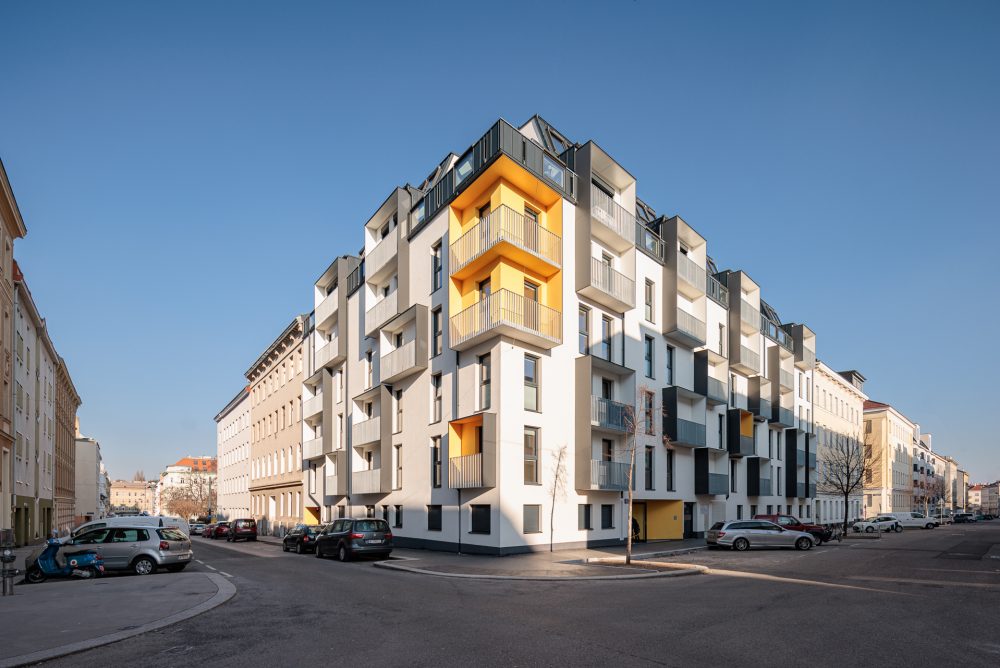 Modernes Wohngebäude mit klaren Linien und gelben Akzenten, Balkone und große Fenster, umgeben von parkenden Autos auf einer Straße.