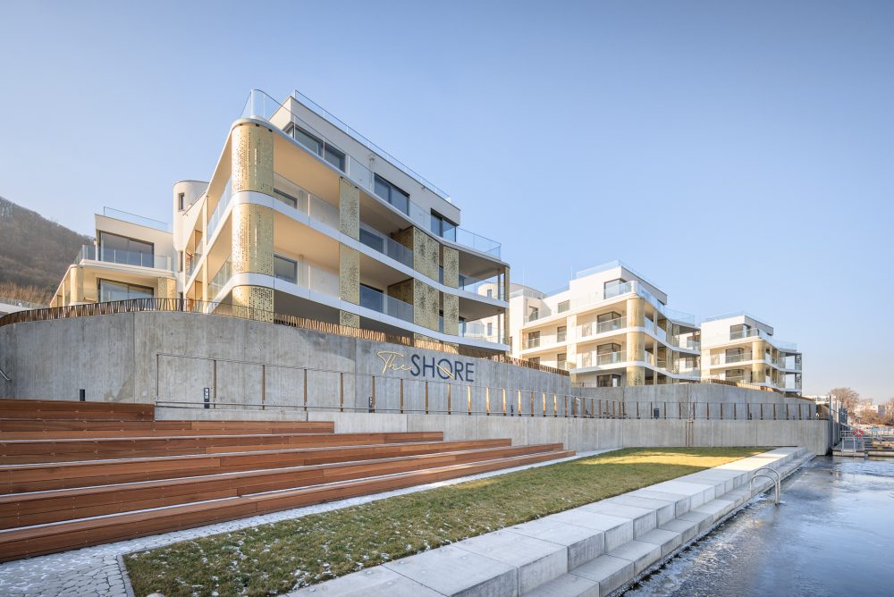 Mehrstöckige moderne Gebäude mit Balkonen und Glasfassaden, davor Treppenstufen und ein Schwimmbecken bei sonnigem Wetter.