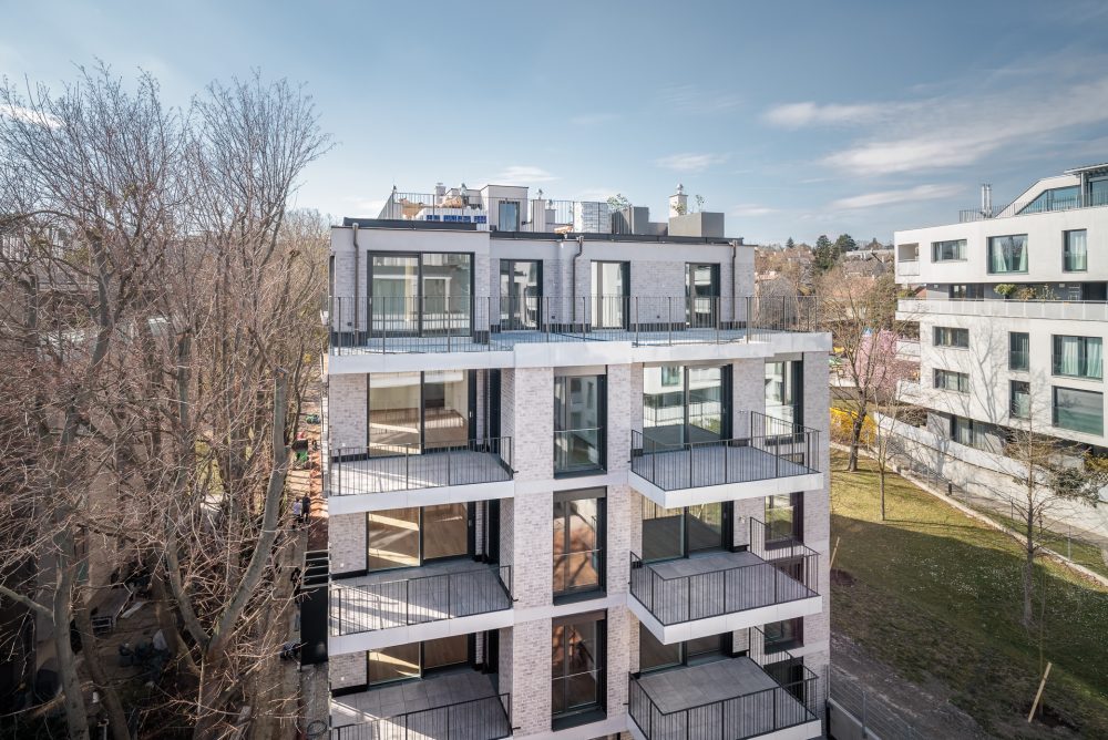 Mehrstöckiges, modernes Gebäude mit großen Fenstern und Balkonen, umgeben von kahlen Bäumen und angrenzenden Wohnhäusern.