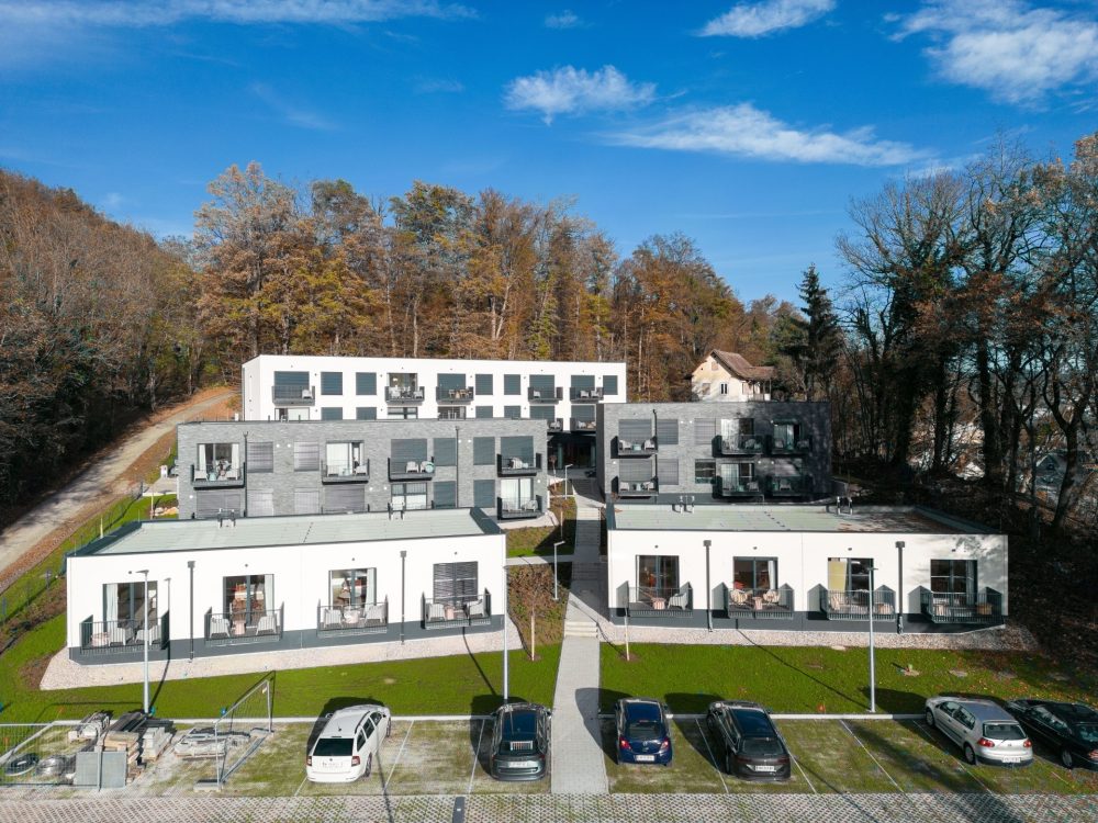 Eine moderne Wohnanlage vor einem bewaldeten Hügel mit drei Gebäuden, Grünflächen und mehreren geparkten Autos im Vordergrund.