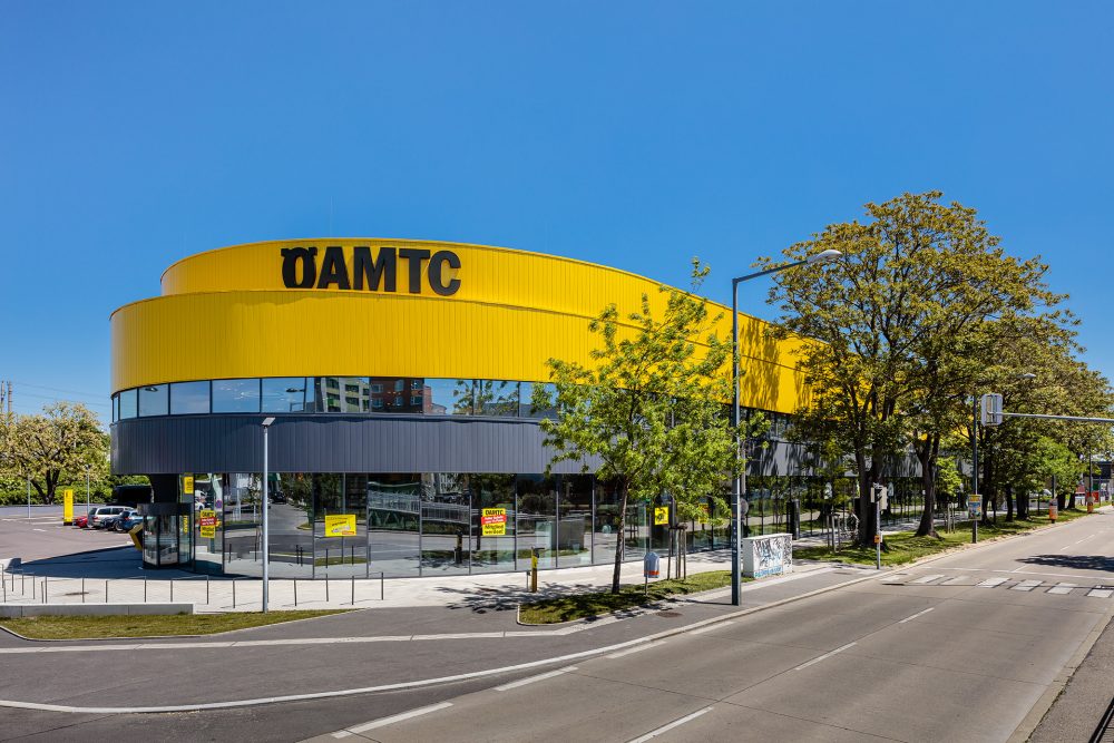 Ein modernes, rundes Gebäude mit gelber und schwarzer Fassade, beschriftet mit ÖAMTC, umgeben von Bäumen an einer Straße.