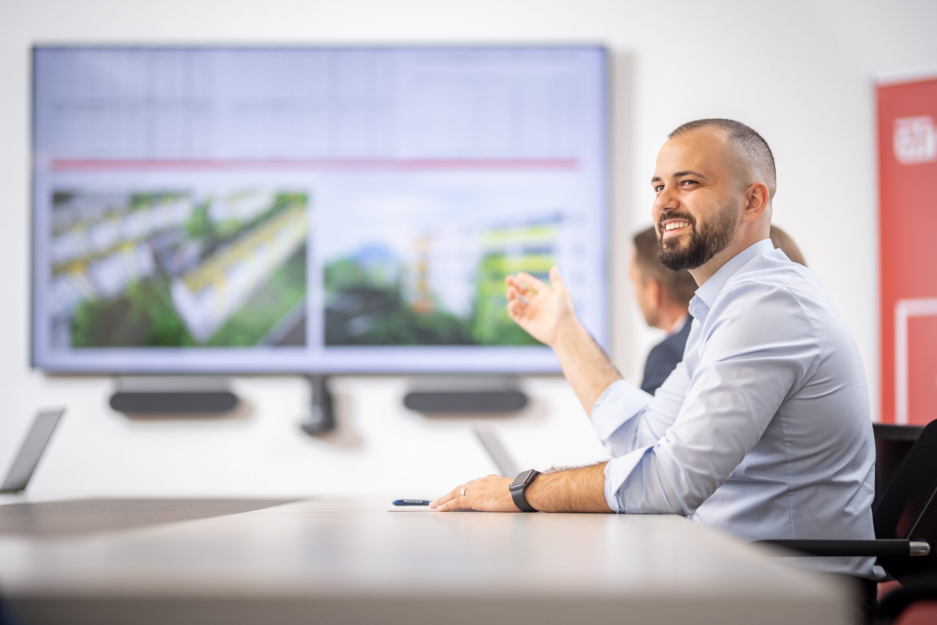 Ein lächelnder Mann im Büro zeigt auf eine große Präsentation auf dem Bildschirm, neben ihm sitzen weitere Personen.