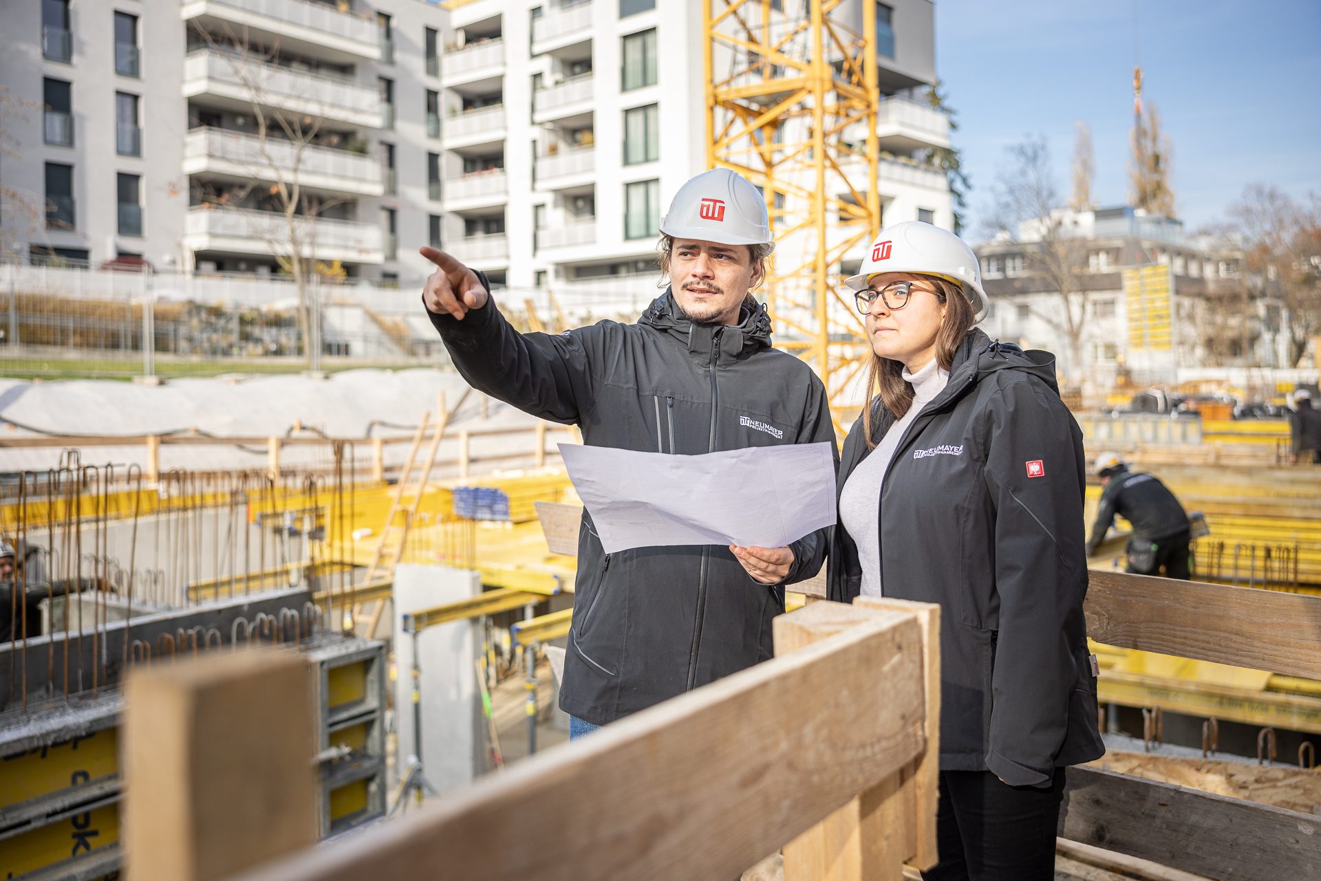 Zwei Personen mit Bauhelmen und Plänen stehen auf einer Baustelle. Eine Person zeigt mit dem Finger, Gebäude sind im Hintergrund.
