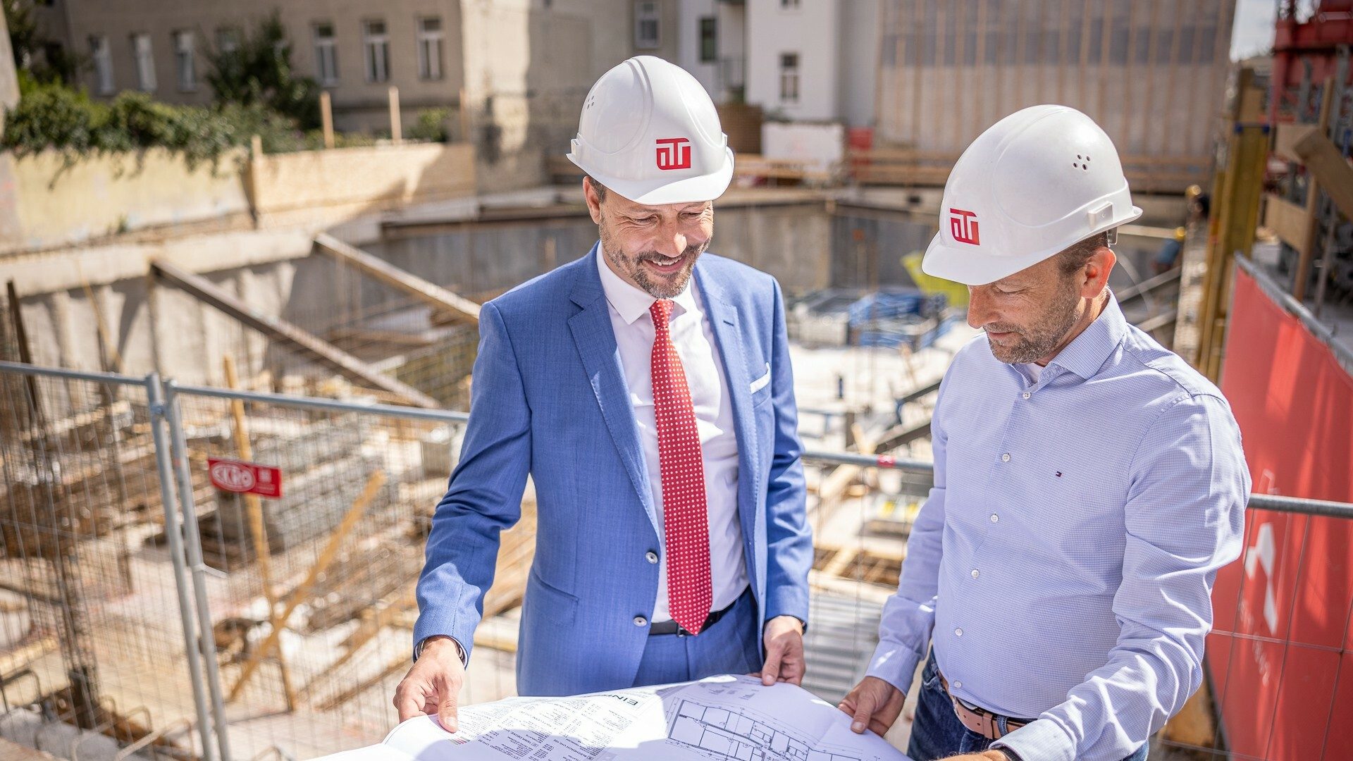 Zwei Männer in Anzügen und weißen Bauhelmen stehen auf einer Baustelle und betrachten einen Bauplan.