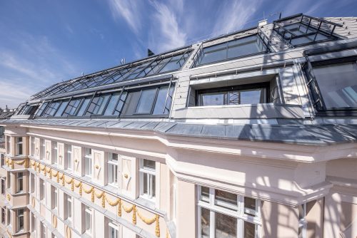 Das Bild zeigt die Fassade eines mehrstöckigen modernen Gebäudes mit großen Dachfenstern und verzierten Elementen, vor einem blauen Himmel.