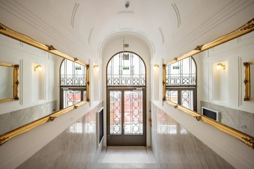 Heller Flur mit hohen Decken, verziert mit goldgerahmten Spiegeln und Wandleuchten; eine Glastür führt nach draußen.