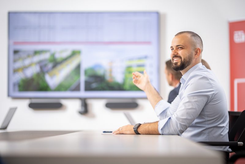 Ein lächelnder Mann im Büro zeigt auf eine große Präsentation auf dem Bildschirm, neben ihm sitzen weitere Personen.