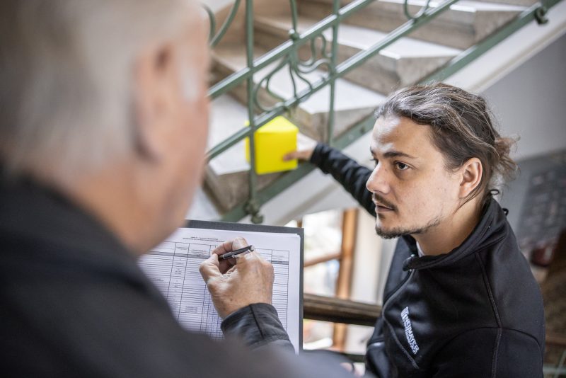 Zwei Männer befinden sich auf einer Treppe, einer schaut nachdenklich, während der andere in einem Klemmbrett schreibt.