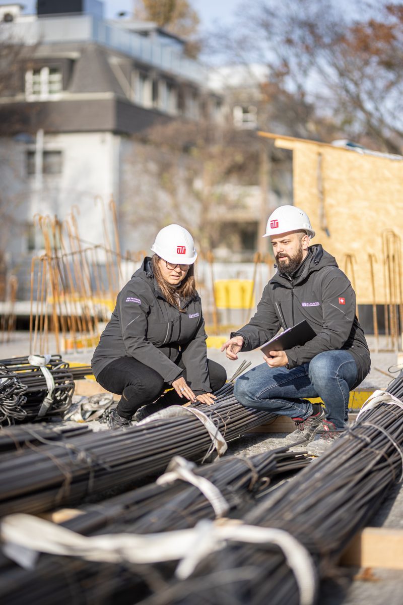 Zwei Personen in Arbeitskleidung und Helmen begutachten Stahlstäbe auf einer Baustelle, während sie miteinander sprechen.
