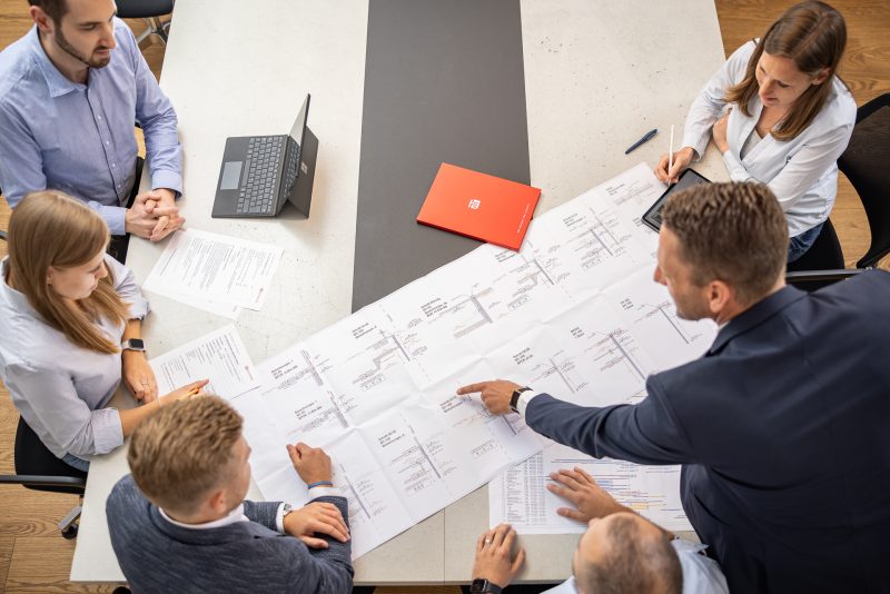 Menschen diskutieren an einem Tisch über Baupläne, umgeben von einem Laptop, Dokumenten und einem roten Ordner.