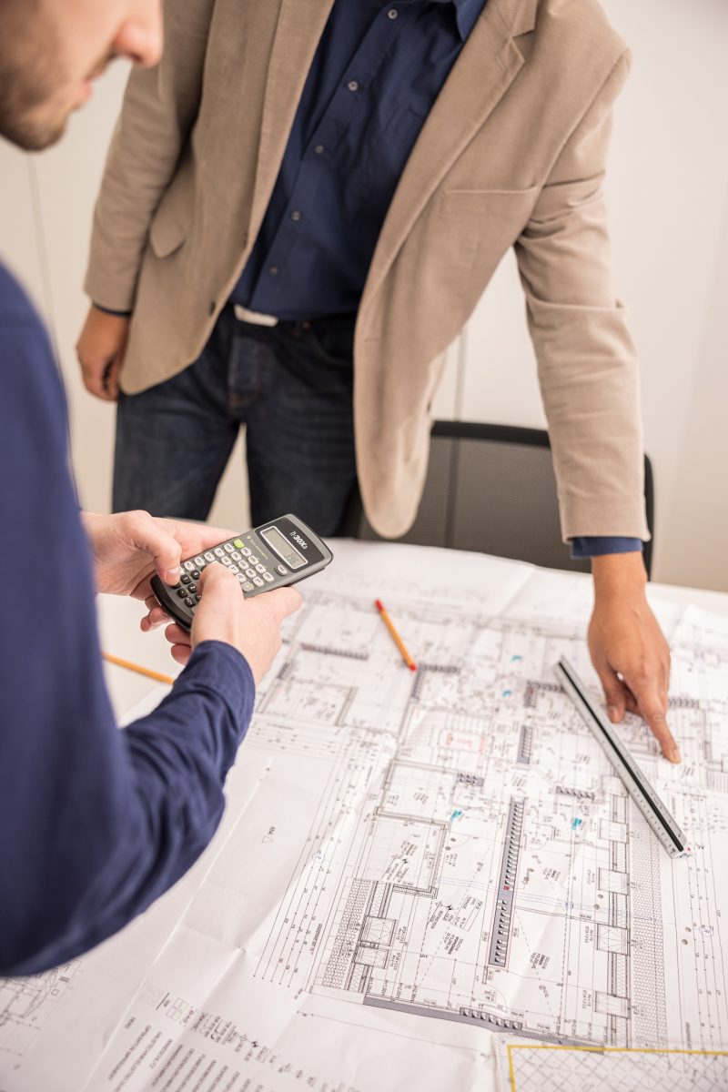 Zwei Männer besprechen Baupläne auf einem Tisch; einer verwendet einen Taschenrechner, der andere zeigt auf den Plan.