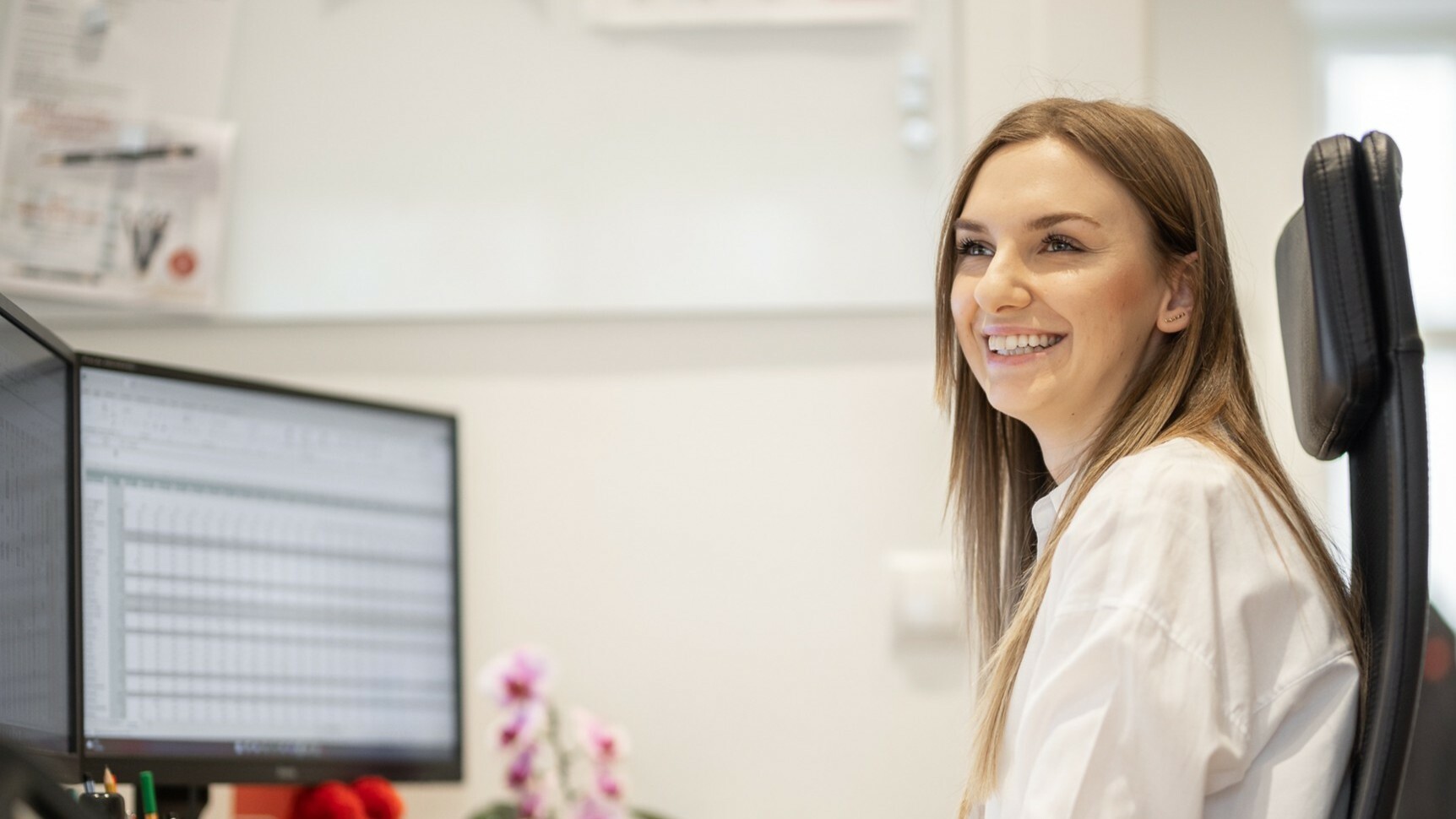 Eine lächelnde Frau sitzt in einem Büro und schaut auf einen Computerbildschirm, auf dem eine Tabelle angezeigt wird.